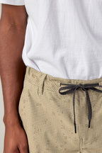 A person wearing a white T-shirt and 686 Men's Everywhere® Pant - Slim Fit in beige with a black drawstring, shown waist to mid-thigh, left arm relaxed by their side.