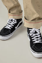 A person stands on a light floor wearing black and white lace-up sneakers and beige 686 Men's Everywhere® Pant - Slim Fit from 686, featuring stain resistance and a red seam at the cuffs.