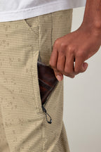 A person wearing 686 Men's Everywhere® Pant - Slim Fit in beige patterned, stain-resistant fabric puts a brown leather wallet into a zippered side pocket, with a white shirt partially visible at the waist.