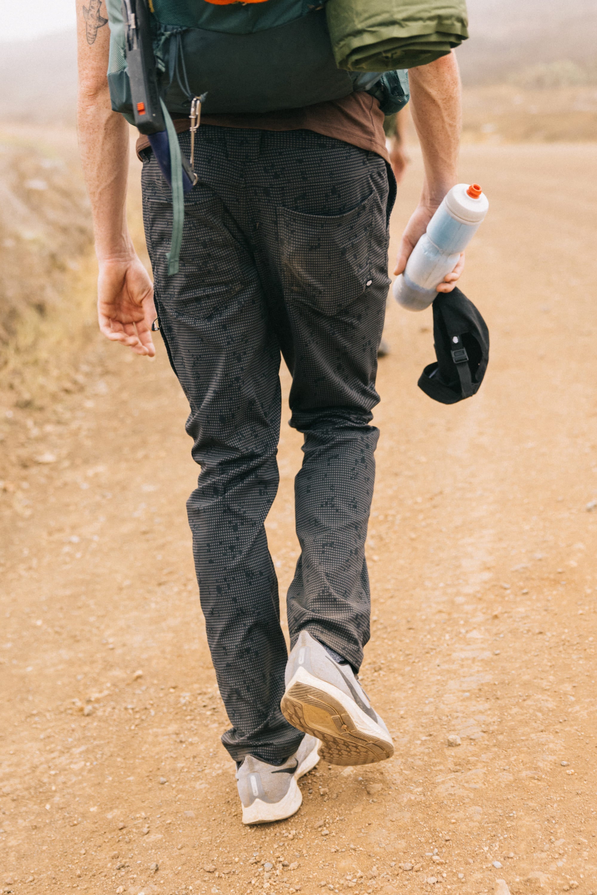 Wearing 686 Men's Everywhere® Pant - Slim Fit, sneakers, and a backpack, a person walks on a dirt path holding a black cap and water bottle in their left hand. The ground-level photo captures them from behind.