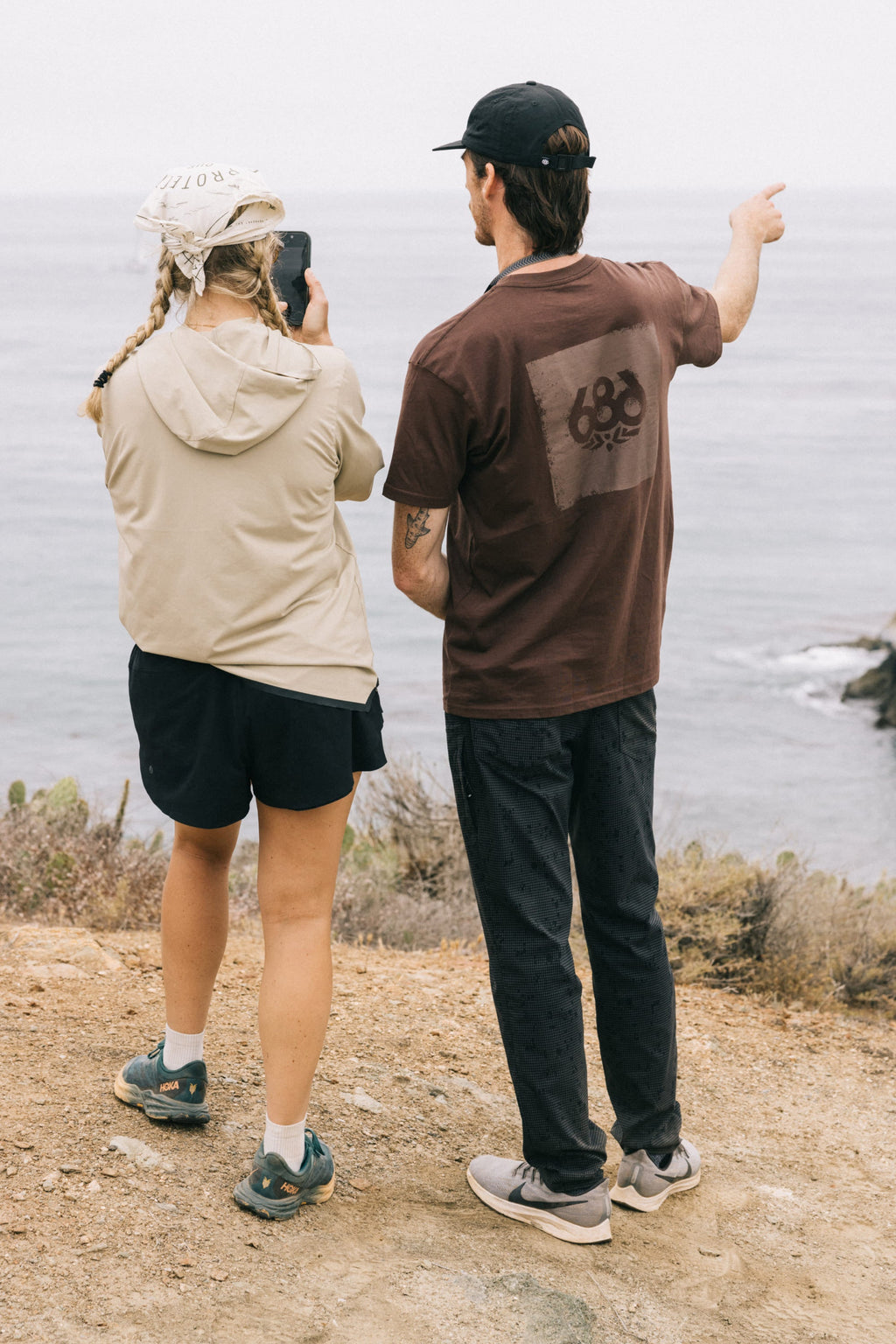 Two people stand on a rocky coastal path, ocean in view. One, in 686 Men's Everywhere® Pant - Slim Fit, snaps a photo while the other points to the water. Both wear casual outdoor outfits and hats, with shrubs and water behind them.