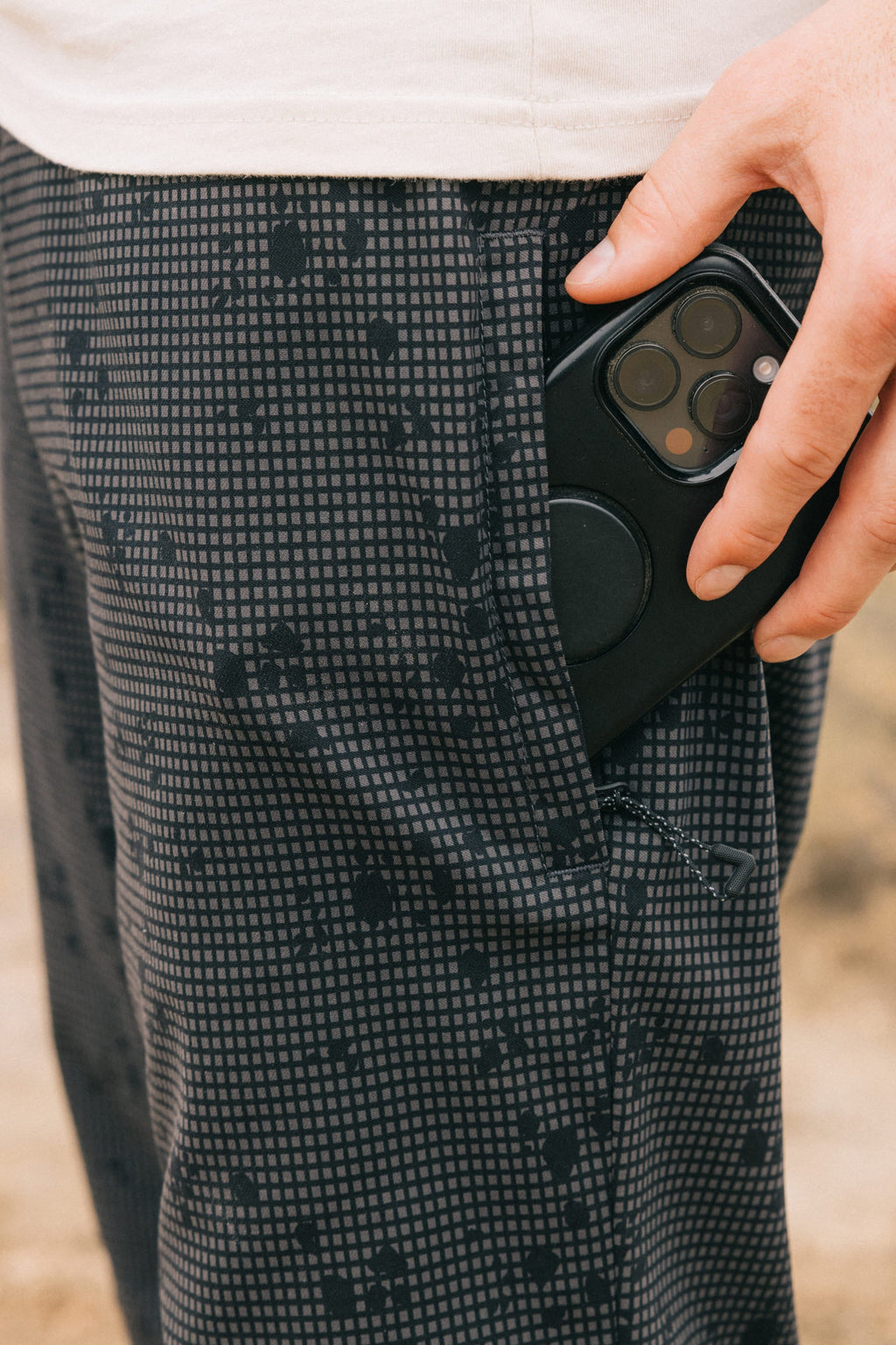A hand places a black smartphone with a circular grip into the pocket of gray, breathable 686 Men's Everywhere® Pant - Slim Fit. The wearer has a white shirt, and the background is blurred.
