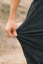 A close-up shows a hand stretching the fabric of 686 Men's Everywhere® Pant - Slim Fit in black and gray check outdoors, highlighting its stretchiness with a blurred dirt or rocky background.