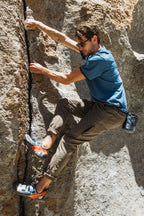 A man in sunglasses, a blue shirt, 686 Men's Everywhere® Pant - Slim Fit by 686, and climbing shoes scales a vertical rocky surface, gripping a crack in the rock.