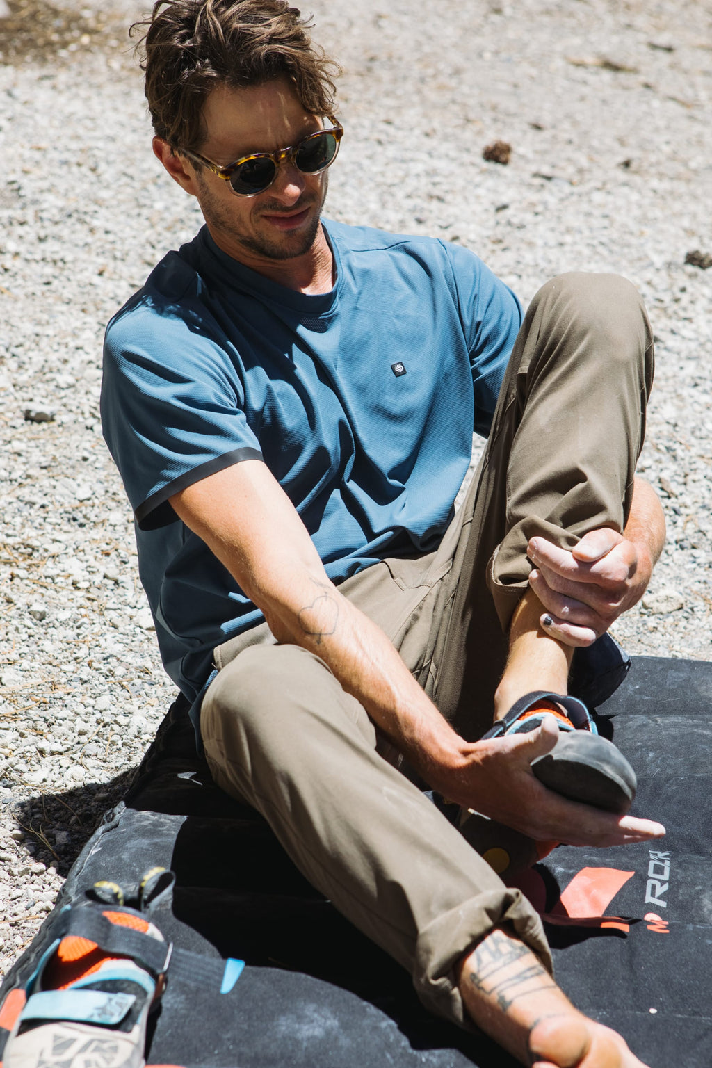 A man in sunglasses sits on a crash pad outdoors, putting on climbing shoes. He wears a blue shirt and breathable pants, with his 686 Men's Everywhere® Pant - Slim Fit by his side, sunlight highlighting his relaxed, focused expression.