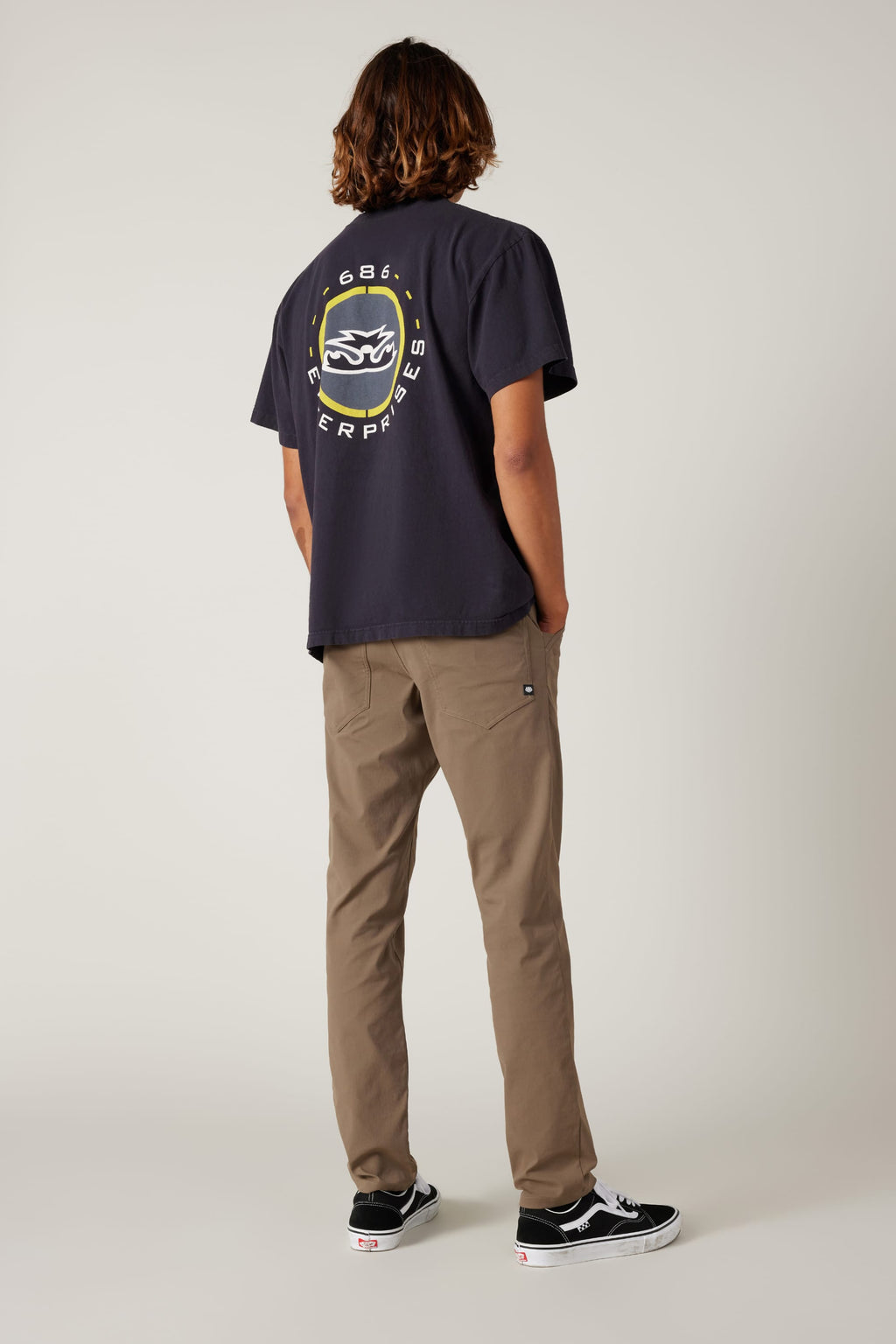 A person stands facing away, wearing a navy T-shirt with a circular white and yellow logo on the back, 686 Men's Everywhere® Pant - Slim Fit in khaki, and black Vans sneakers with white soles, set against a plain light background.