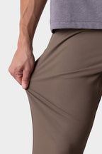 A person wearing 686 Men's Everywhere® Pant - Slim Fit in taupe and a gray shirt stretches the fabric with their left hand, showing the flexibility and breathability of these 686 pants.