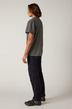 A person with medium-length brown hair stands in profile left, wearing a gray T-shirt, 686 Men's Anything Cargo Pant - Relaxed Fit by 686 with 13 pockets and 4-way stretch, plus black sneakers, against a plain light background.