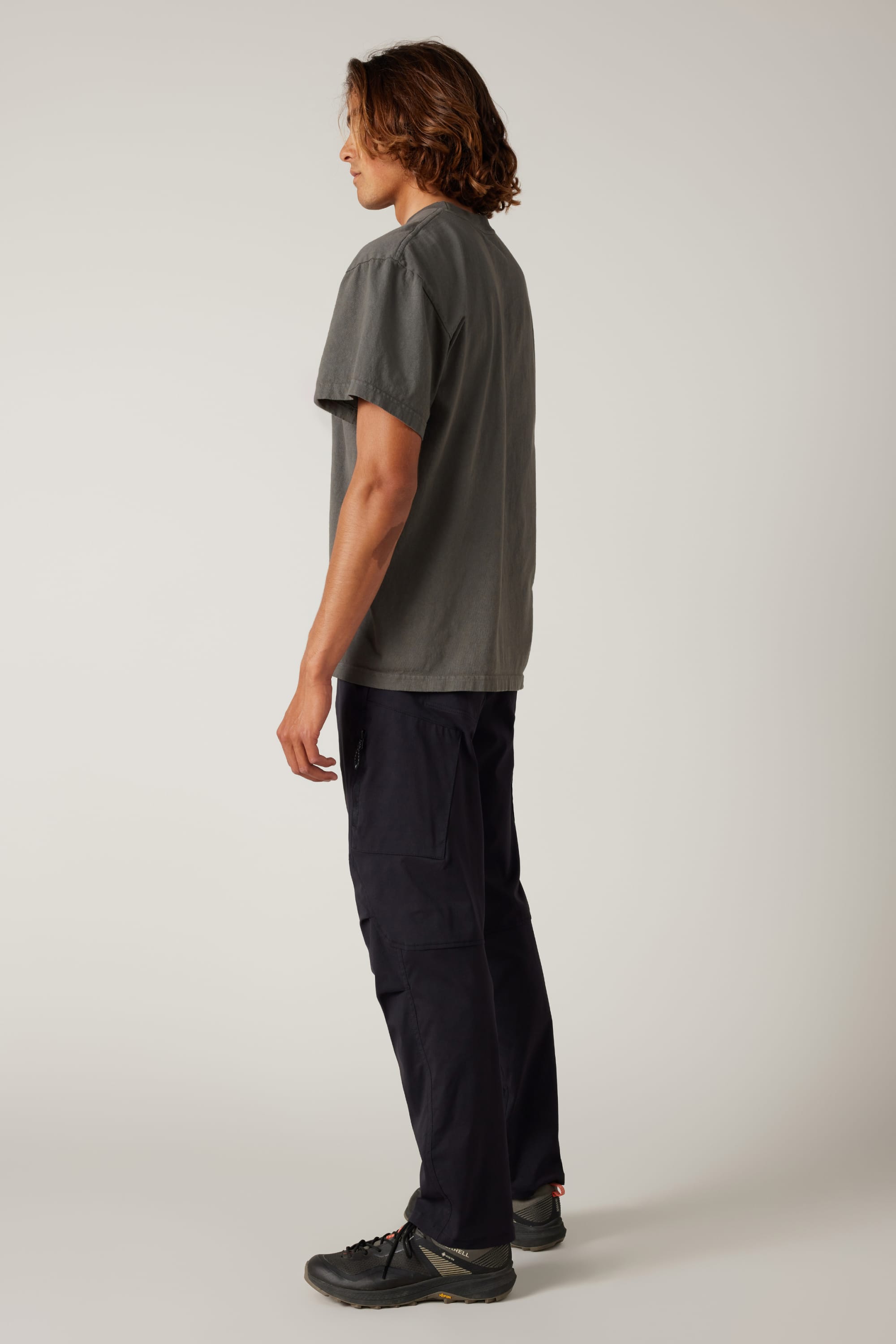 A person with medium-length brown hair stands in profile left, wearing a gray T-shirt, 686 Men's Anything Cargo Pant - Relaxed Fit by 686 with 13 pockets and 4-way stretch, plus black sneakers, against a plain light background.