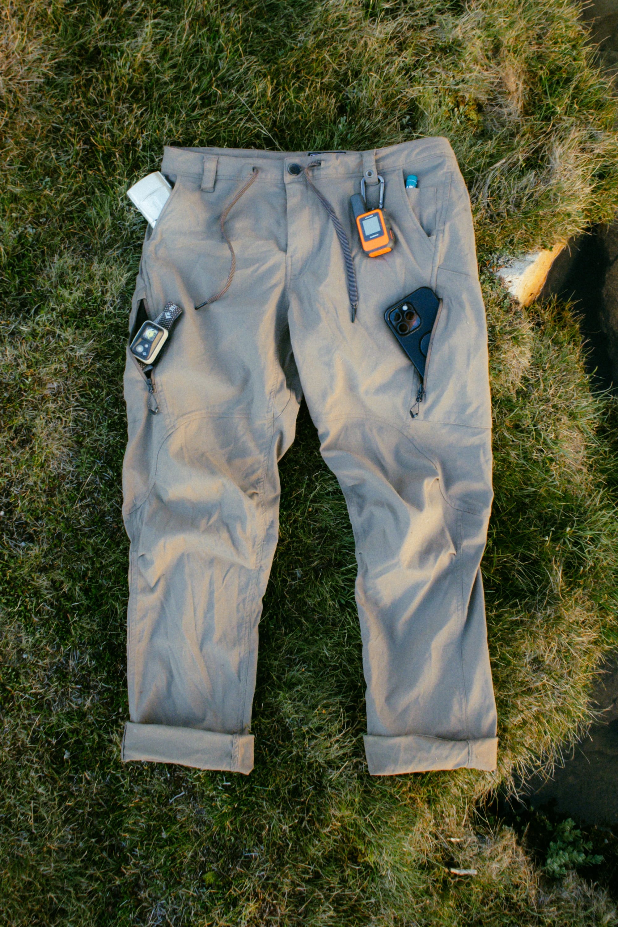 A pair of 686 Men's Anything Cargo Pant - Relaxed Fit in light brown lies on the grass, with a GPS, smartphone, walkie-talkie, and folded map tucked into its spacious 13 pockets.