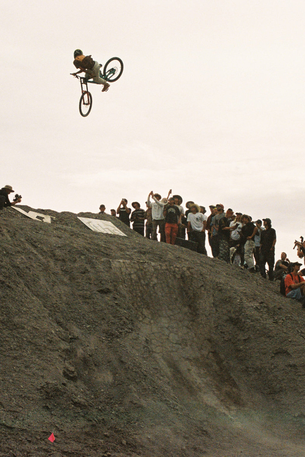 A cyclist launches off a dirt ramp before a cheering crowd, wearing 686 Men's Anything Cargo Pant - Relaxed Fit by 686. The overcast sky adds energy to the lively scene as onlookers capture the action.