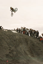 A cyclist launches off a dirt ramp before a cheering crowd, wearing 686 Men's Anything Cargo Pant - Relaxed Fit by 686. The overcast sky adds energy to the lively scene as onlookers capture the action.