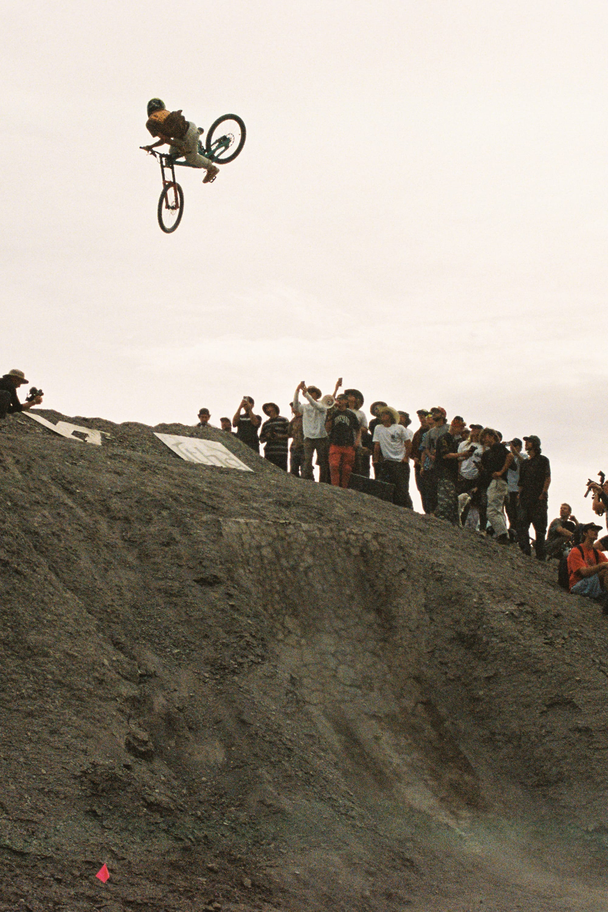 A cyclist launches off a dirt ramp before a cheering crowd, wearing 686 Men's Anything Cargo Pant - Relaxed Fit by 686. The overcast sky adds energy to the lively scene as onlookers capture the action.