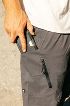 A person in a white shirt wears 686 Men's Anything Cargo Pant - Relaxed Fit in gray and puts a black smartphone into the zippered pocket. The outdoor photo shows the 4-way stretch technical pants by 686 in sunlight.