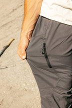 A person wearing 686 Men's Anything Cargo Pant - Relaxed Fit in gray with a zippered pocket stands on sandy ground, hand in pocket. Only part of their torso and arm are visible.