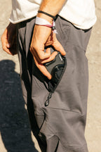 Wearing the 686 Men's Anything Cargo Pant - Relaxed Fit and a white shirt, a person places a compact Canon camera into a zippered side pocket. They have a black hair tie, a pink fabric bracelet, and stand on sunlit pavement.