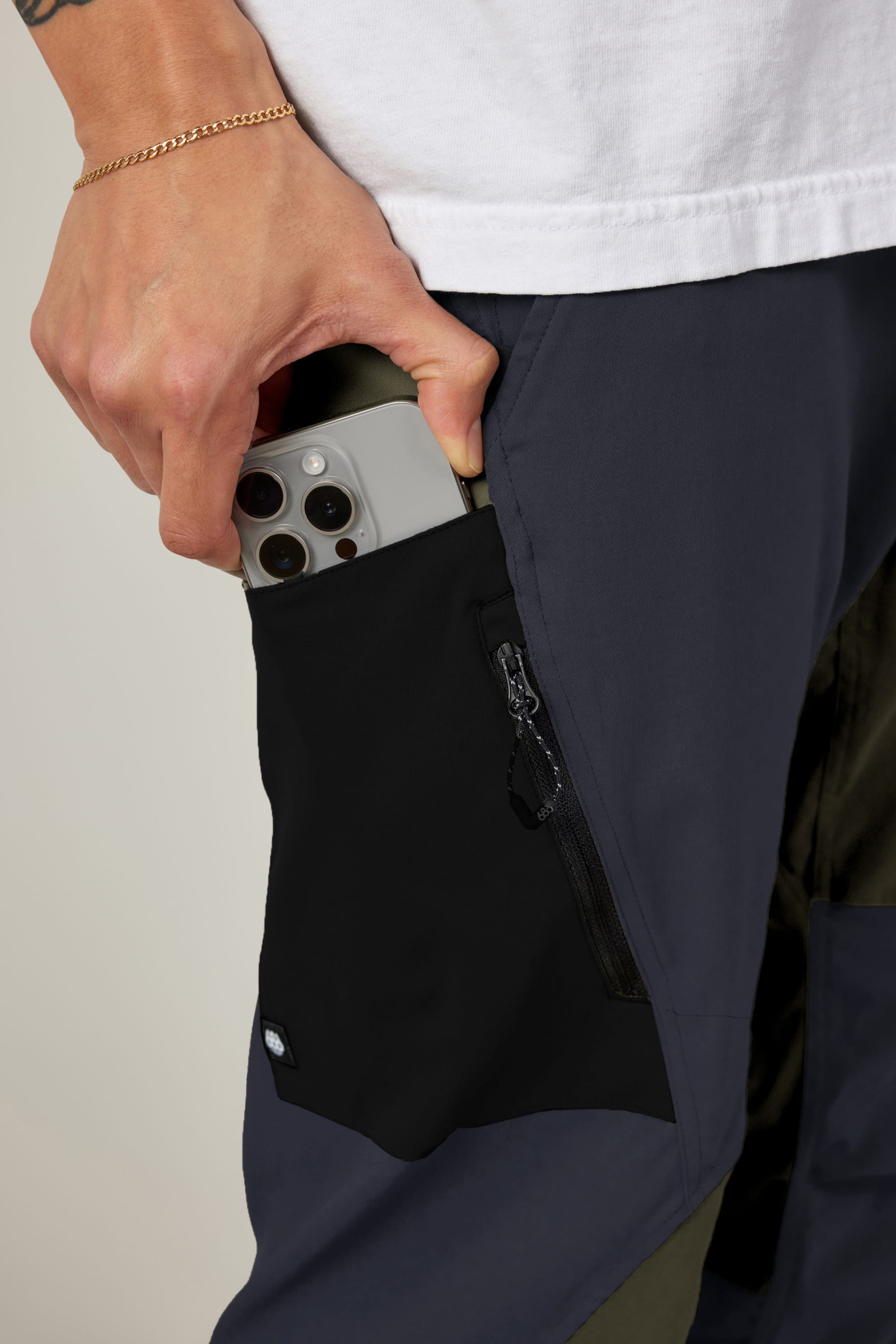 Someone wearing a white shirt and 686 Men's Anything Cargo Pant - Relaxed Fit in dark color slips a triple-lens smartphone into a zippered side pocket. A thin chain bracelet is partly visible on their hand.