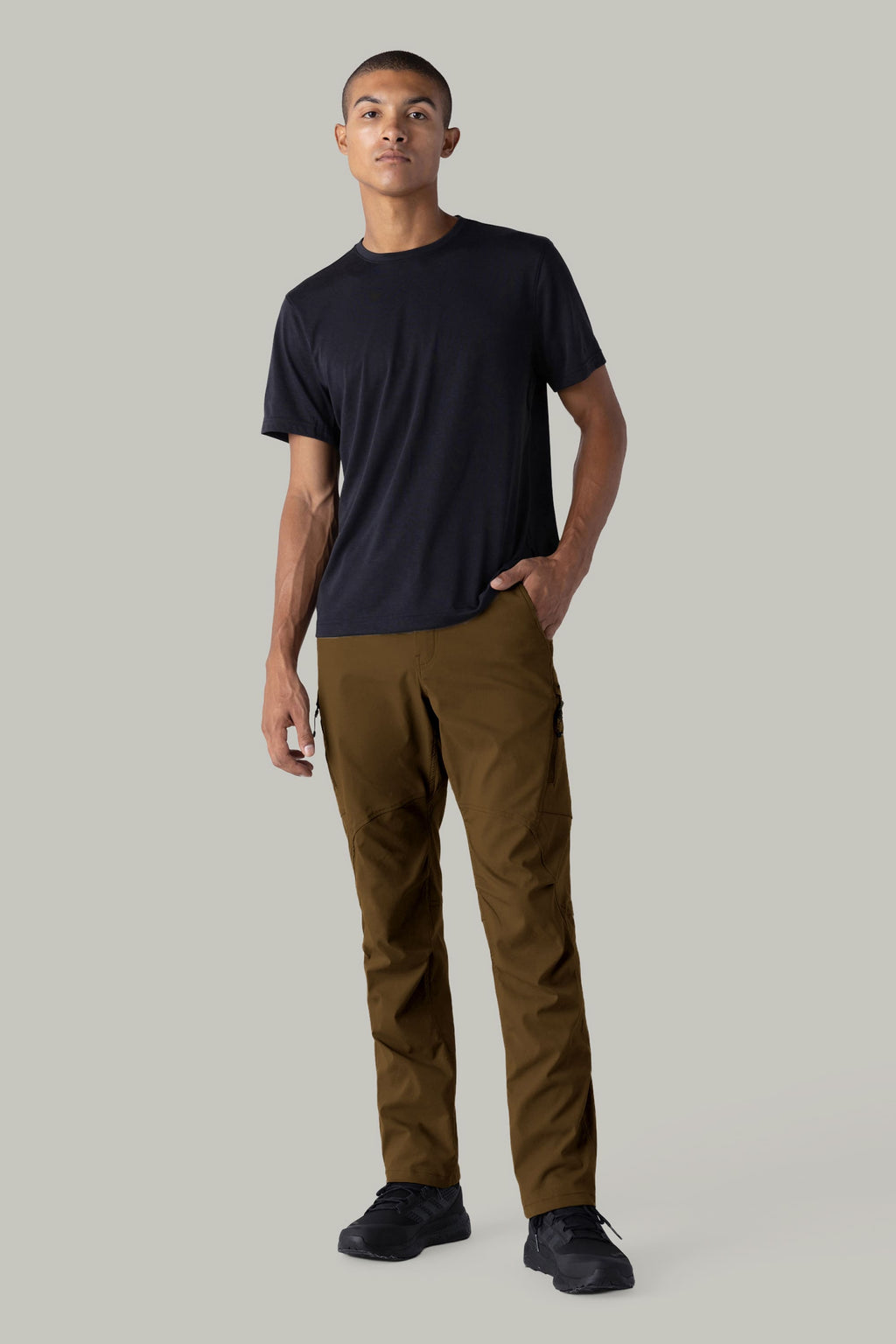 A person stands against a plain gray background wearing a black t-shirt, 686 Men's Anything Cargo Pant - Relaxed Fit in brown, and black shoes, with one hand in their pocket and a neutral expression.