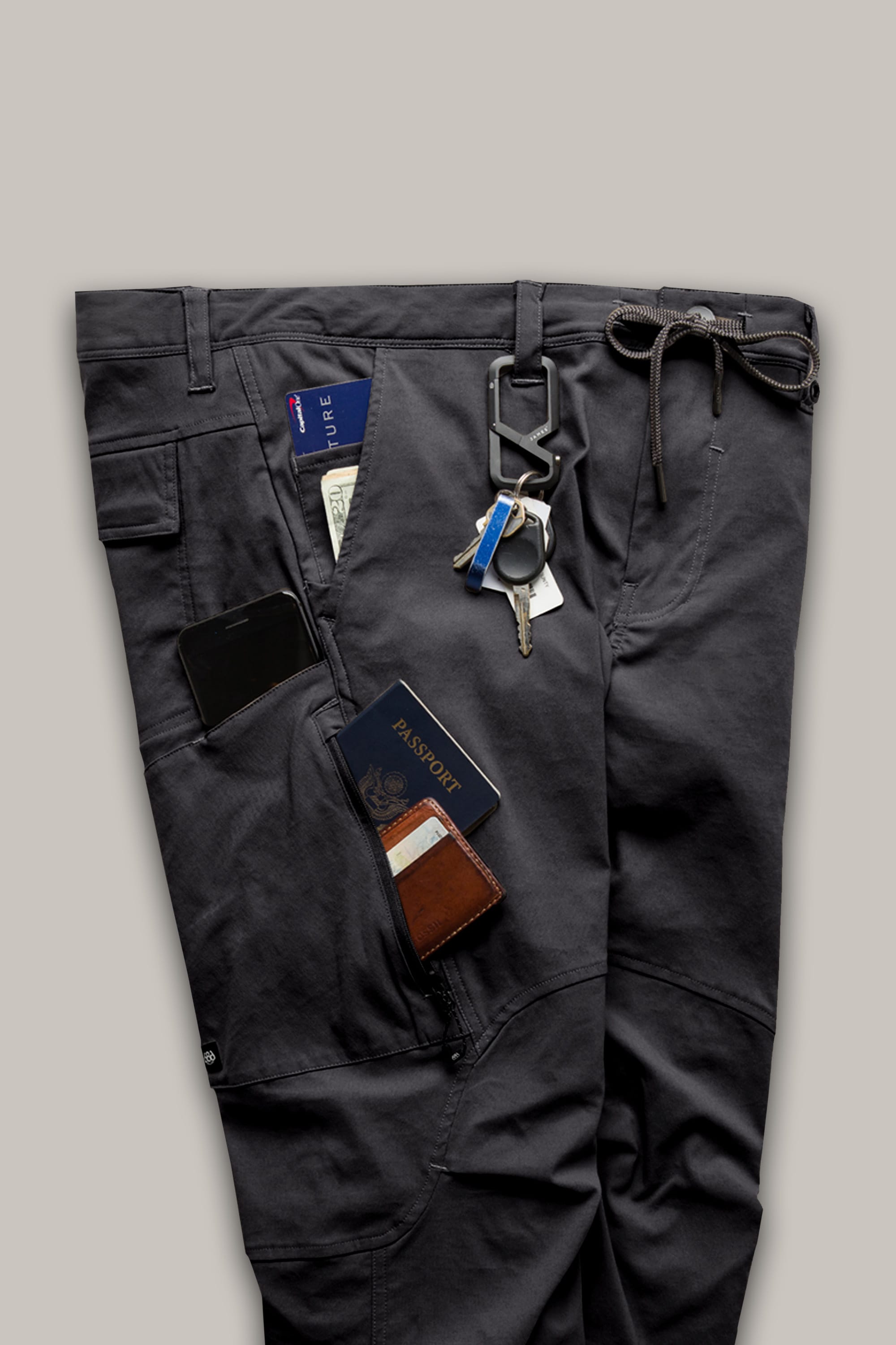 The 686 Men's Anything Cargo Pant - Relaxed Fit by 686 is shown laid flat in dark gray with 13 pockets holding a passport, wallet, smartphone, folded cash, and keys clipped to a belt loop, highlighting its organized, 4-way stretch design.