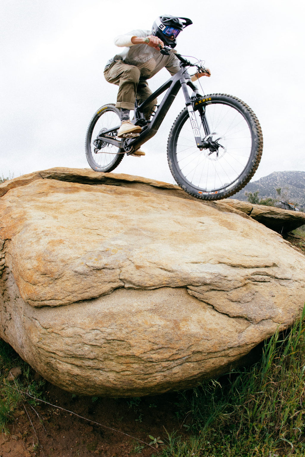 A mountain biker in the 686 Men's Anything Cargo Pant - Relaxed Fit rides over a large rock on a rugged trail, wheels airborne and challenging terrain below.