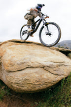 A mountain biker in the 686 Men's Anything Cargo Pant - Relaxed Fit rides over a large rock on a rugged trail, wheels airborne and challenging terrain below.