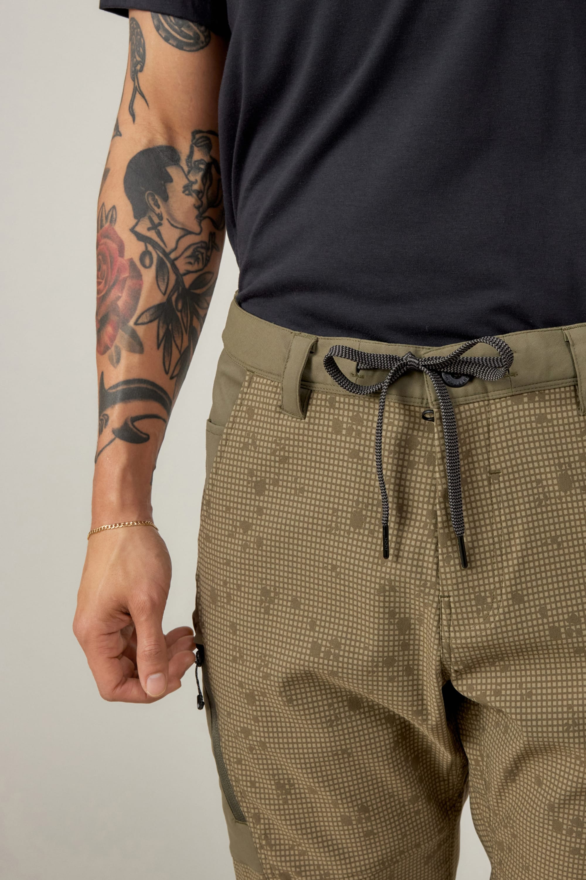 A person wears a black t-shirt and 686 Men's Anything Cargo Pant - Relaxed Fit in olive green, styled with a black-and-white cord belt. Their tattooed left arm with a thin bracelet is visible.
