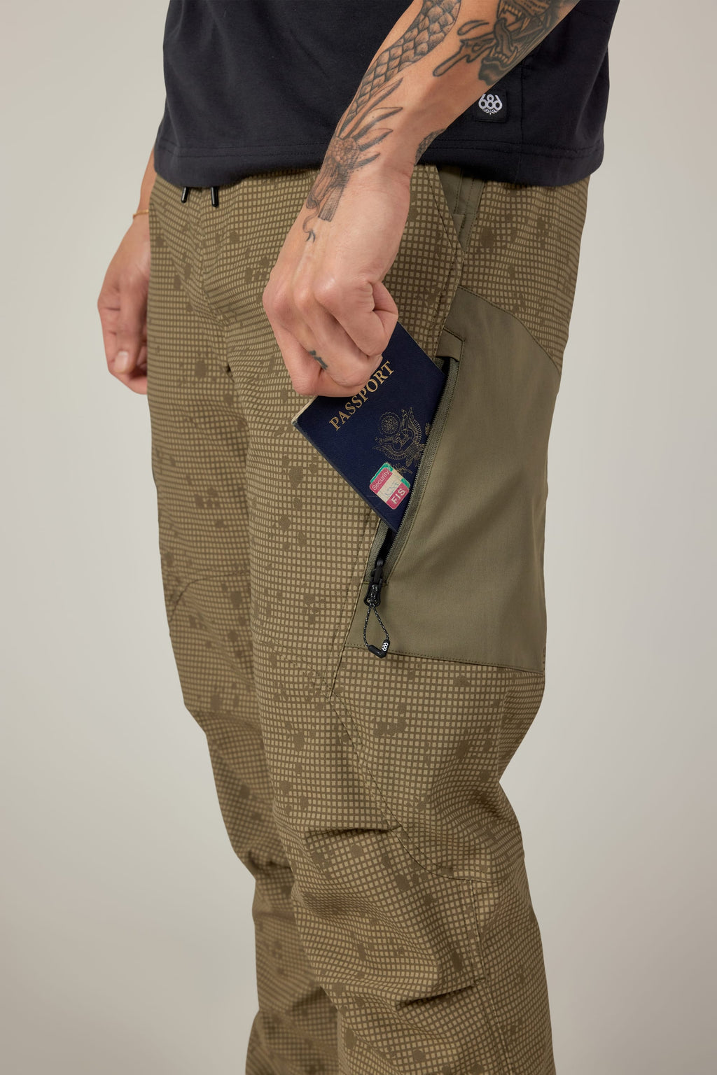 A person with tattooed arms slides a U.S. passport into the zippered thigh pocket of 686 Men's Anything Cargo Pant - Relaxed Fit, a 13-pocket, 4-way stretch olive green pant, paired with a black shirt—only their torso and legs are visible.