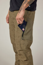 A person with tattooed arms slides a U.S. passport into the zippered thigh pocket of 686 Men's Anything Cargo Pant - Relaxed Fit, a 13-pocket, 4-way stretch olive green pant, paired with a black shirt—only their torso and legs are visible.
