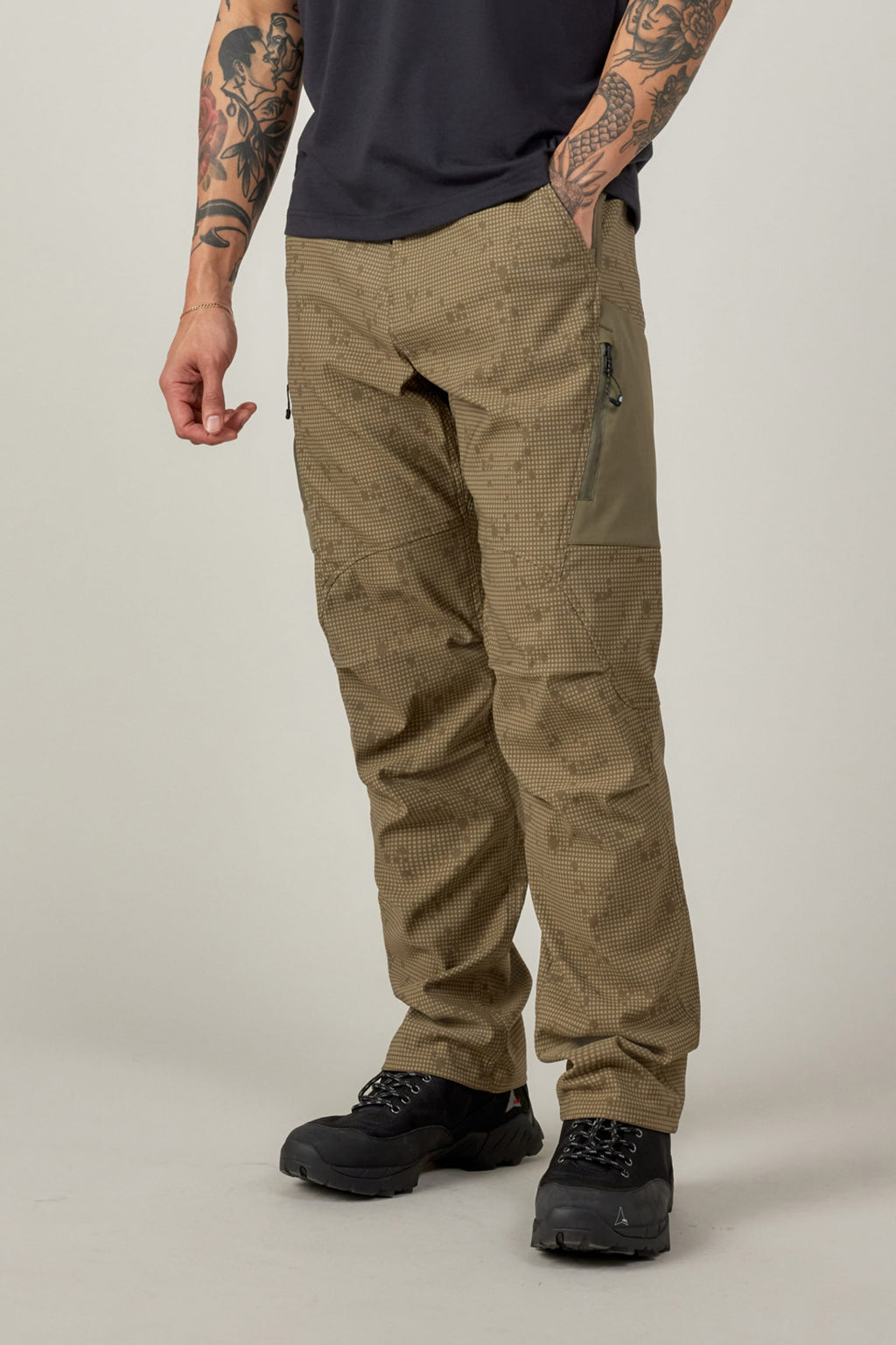 A person wears 686 Men's Anything Cargo Pant - Relaxed Fit in tan with a pattern, paired with a dark t-shirt and black shoes. Both arms are tattooed, one hand is in a pocket, and the plain background is light-colored.