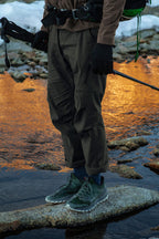 Wearing green trail shoes, gloves, and holding trekking poles, a person in 686 Men's Anything Cargo Pant - Relaxed Fit stands on a rock by a stream with snow and sunset in the background.