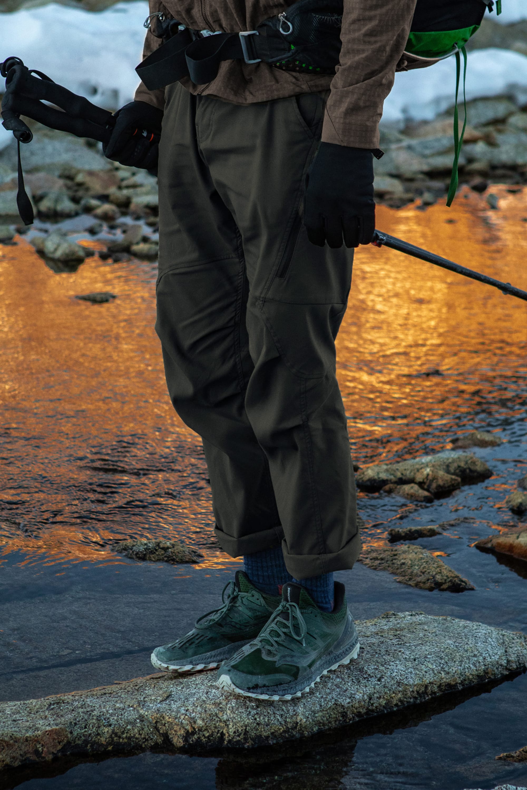 Wearing green trail shoes, gloves, and holding trekking poles, a person in 686 Men's Anything Cargo Pant - Relaxed Fit stands on a rock by a stream with snow and sunset in the background.