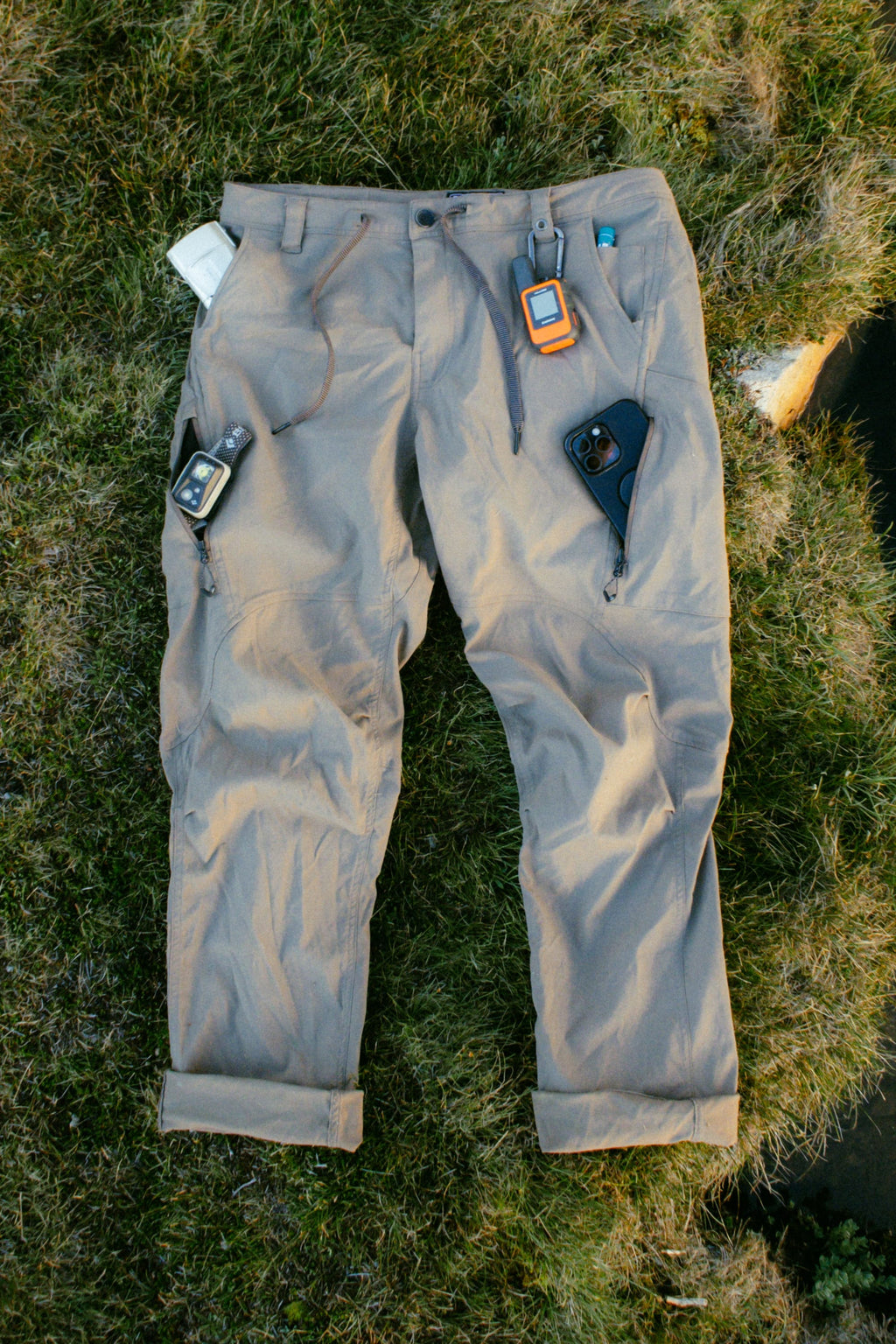 The 686 Men's Anything Cargo Pant - Relaxed Fit by 686 is shown on grass with a smartphone, GPS device, and small electronics organized in its multiple 4-way stretch pockets and waistband.