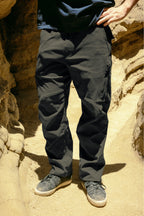 A person in 686 Men's Anything Cargo Pant - Relaxed Fit by 686 and gray sneakers stands on sandy ground between rocky walls, hands on hips. Upper body is partially out of frame.