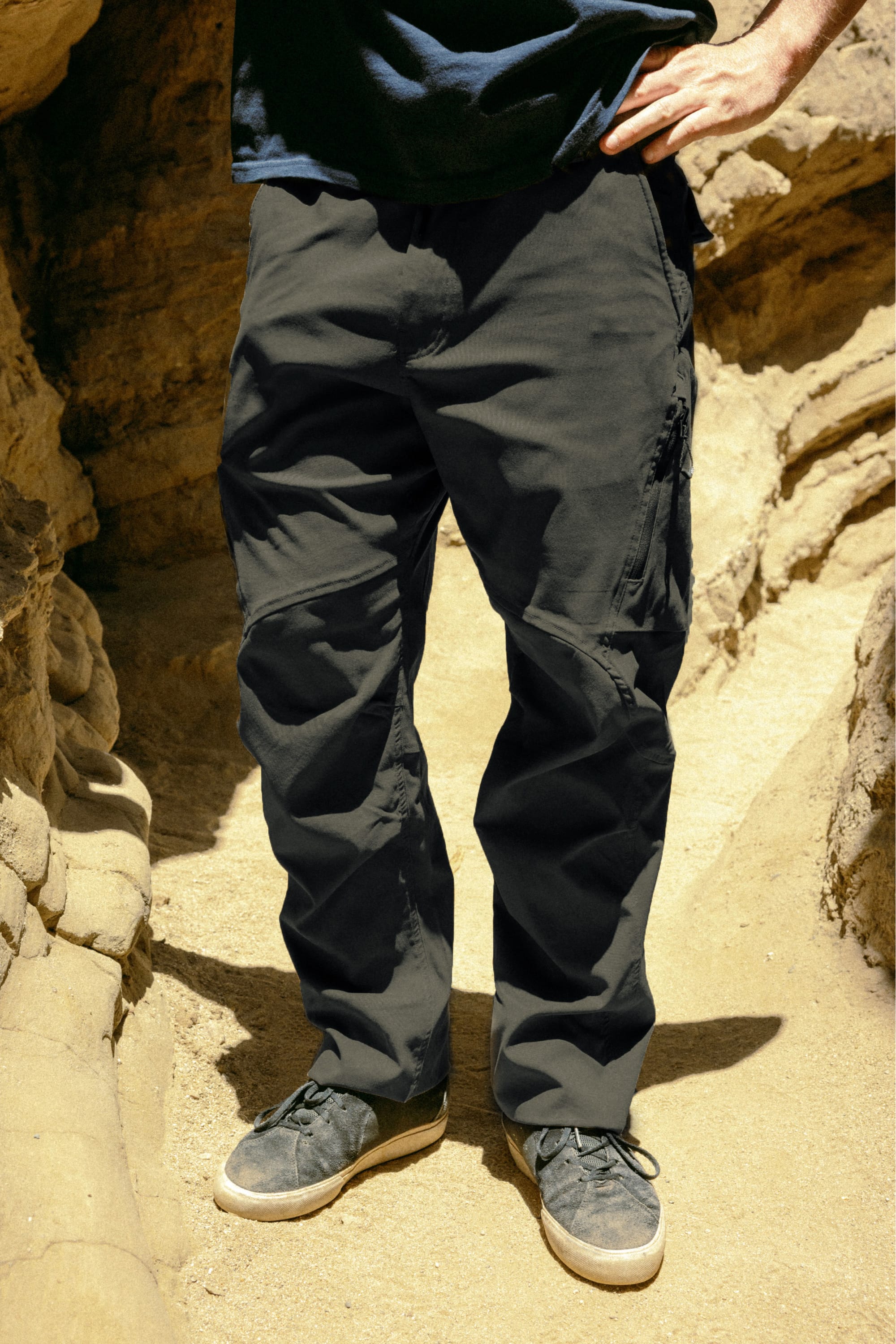 A person in 686 Men's Anything Cargo Pant - Relaxed Fit by 686 and gray sneakers stands on sandy ground between rocky walls, hands on hips. Upper body is partially out of frame.