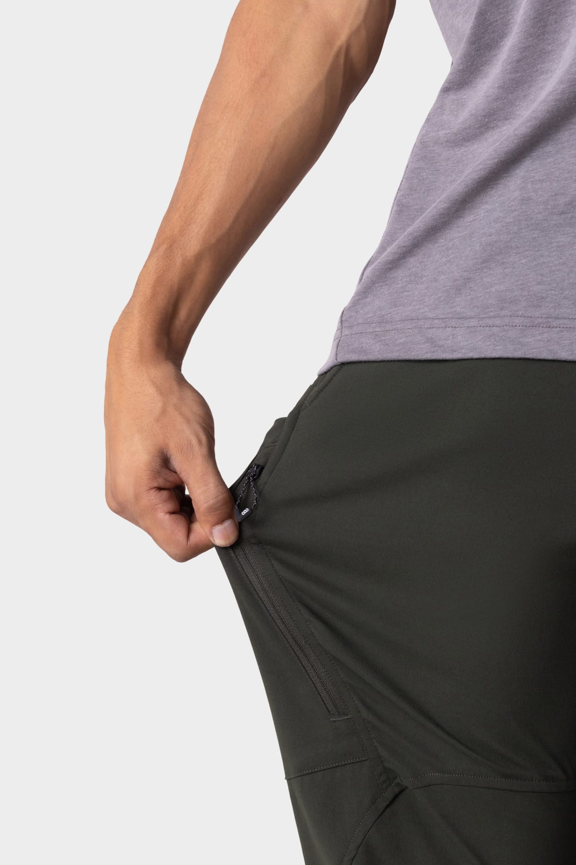 A person wearing the 686 Men's Anything Cargo Pant - Relaxed Fit in dark green pulls open the side zipper pocket, highlighting its 4-way stretch and functional pocket features.