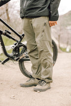 Wearing 686 Men's Anything Cargo Pant - Relaxed Fit, a person stands beside a mountain bike on a dirt trail, showing only their lower half in the olive green technical cargo pants and hiking shoes.