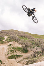 A mountain biker in a helmet and 686 Men's Anything Cargo Pant - Relaxed Fit soars mid-air over a dirt trail, surrounded by grassy hills and leafless trees under a cloudy sky.