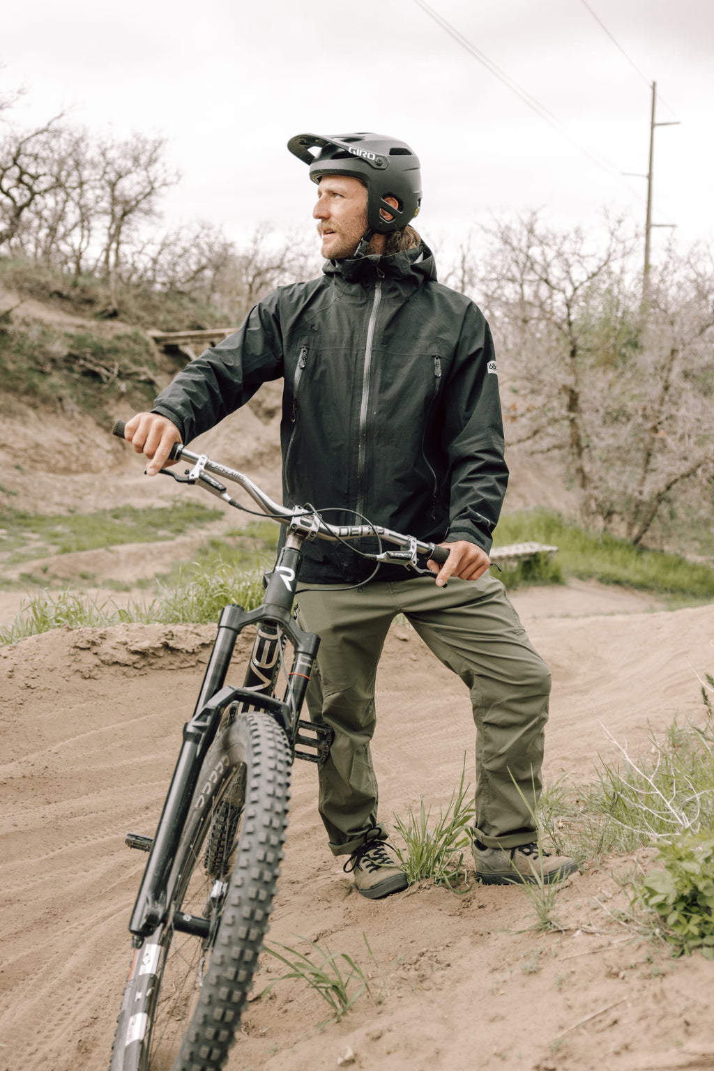 A man in a helmet and outdoor gear, featuring 686 Men's Anything Cargo Pant - Relaxed Fit with 13 pockets, stands beside a mountain bike on a dirt trail. Leafless trees and an overcast sky complete the backdrop.