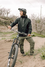 A man in a helmet and outdoor gear, featuring 686 Men's Anything Cargo Pant - Relaxed Fit with 13 pockets, stands beside a mountain bike on a dirt trail. Leafless trees and an overcast sky complete the backdrop.