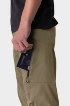 A person wearing the 686 Men's Anything Cargo Pant - Relaxed Fit in olive green slides a dark blue passport halfway into the zippered side pocket, while also wearing a black shirt.