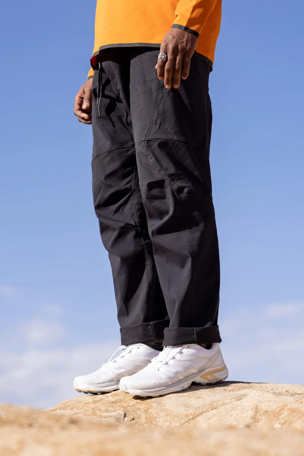 Against a clear blue sky, only the lower half of a person is visible, standing on a rock in 686 Men's Anything Cargo Pant - Relaxed Fit (black) by 686, paired with white sneakers and a bright orange shirt.