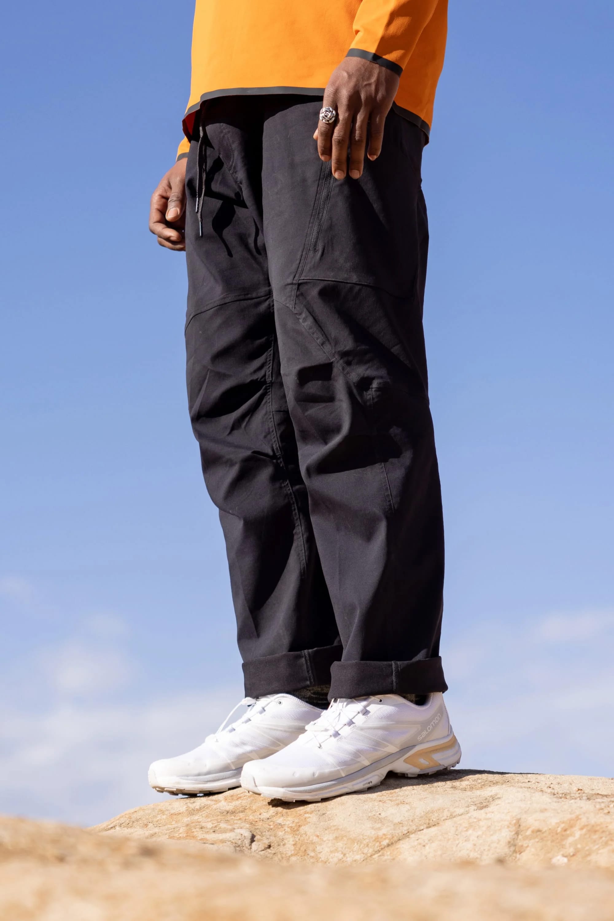 Against a clear blue sky, only the lower half of a person is visible, standing on a rock in 686 Men's Anything Cargo Pant - Relaxed Fit (black) by 686, paired with white sneakers and a bright orange shirt.