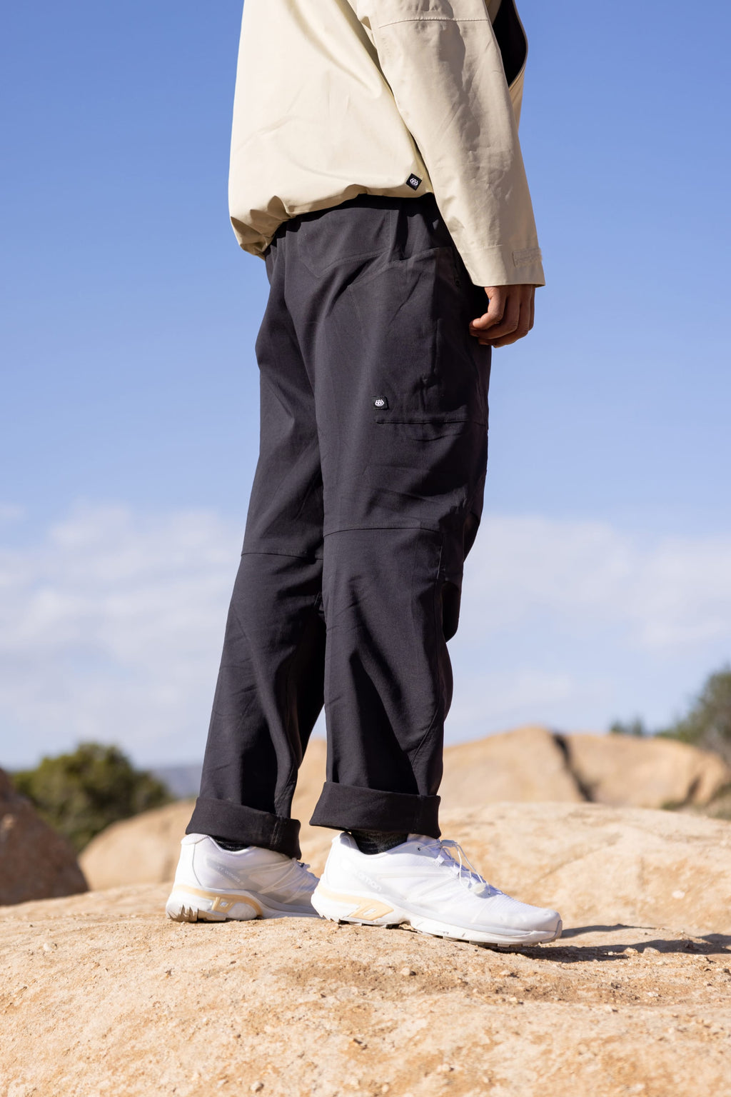 A person stands on rocks outdoors, showing only their lower half, wearing 686 Men's Anything Cargo Pant - Relaxed Fit in black, white sneakers, and a beige jacket. The sky and distant trees appear in the background.