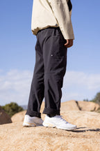 A person stands on rocks outdoors, showing only their lower half, wearing 686 Men's Anything Cargo Pant - Relaxed Fit in black, white sneakers, and a beige jacket. The sky and distant trees appear in the background.