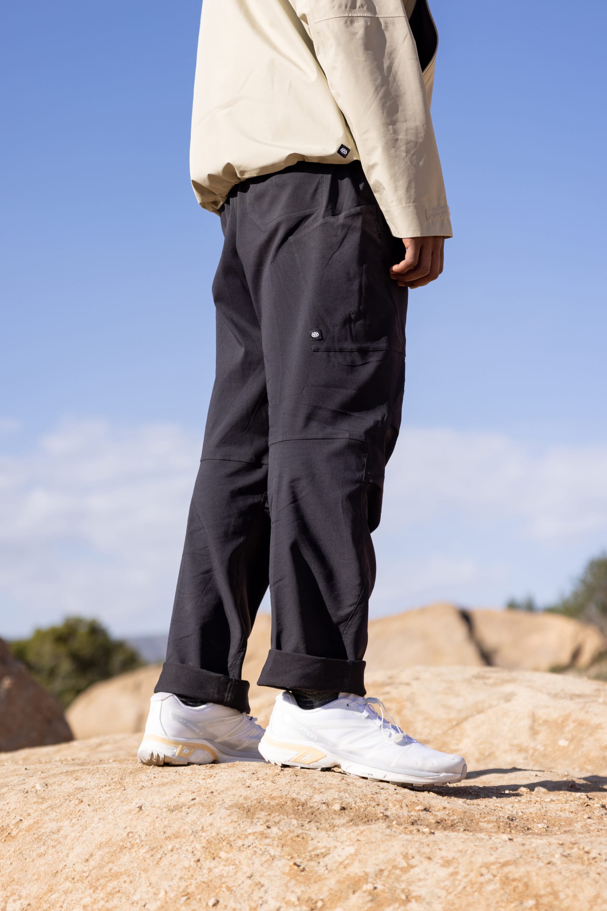 A person stands on rocks outdoors, showing only their lower half, wearing 686 Men's Anything Cargo Pant - Relaxed Fit in black, white sneakers, and a beige jacket. The sky and distant trees appear in the background.