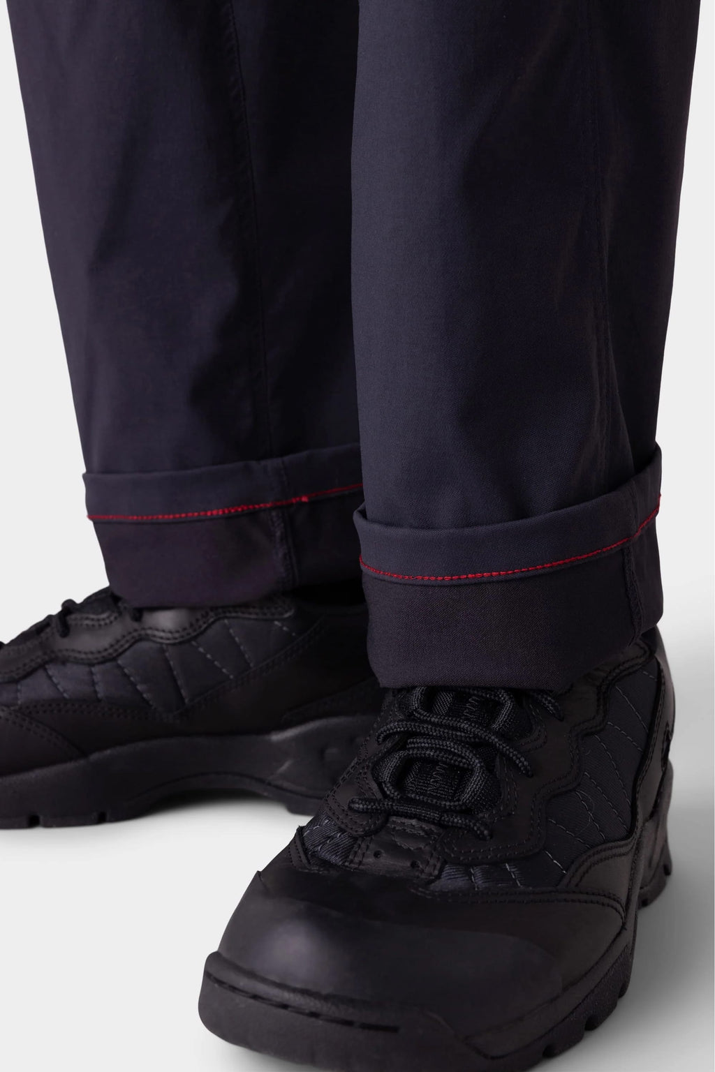 Close-up of a person wearing 686 Men's Anything Cargo Pant - Relaxed Fit by 686 in black, featuring red cuff stitching and paired with black lace-up athletic shoes. The pants are slightly rolled up to highlight the red stitching detail.
