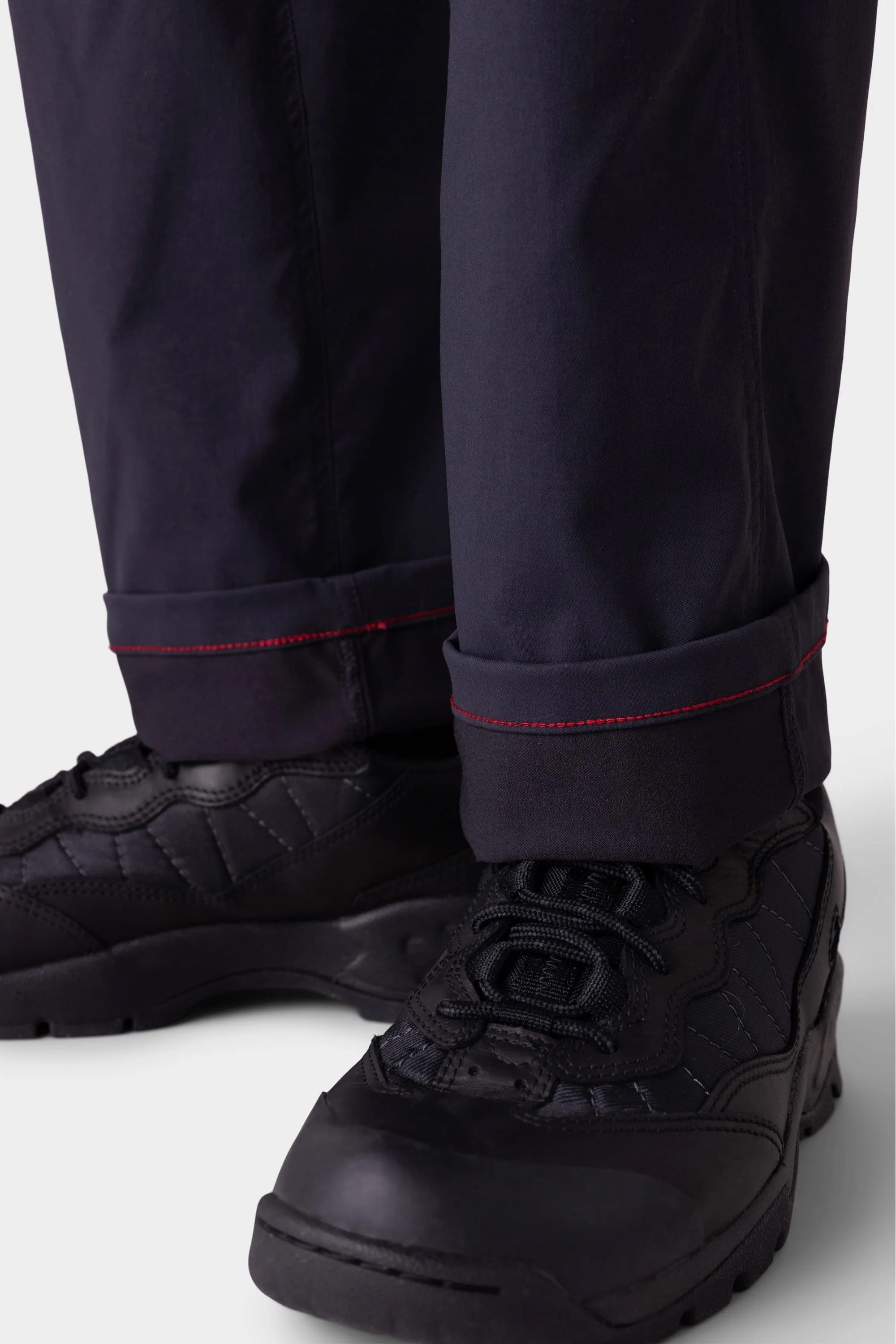 Close-up of a person wearing 686 Men's Anything Cargo Pant - Relaxed Fit by 686 in black, featuring red cuff stitching and paired with black lace-up athletic shoes. The pants are slightly rolled up to highlight the red stitching detail.