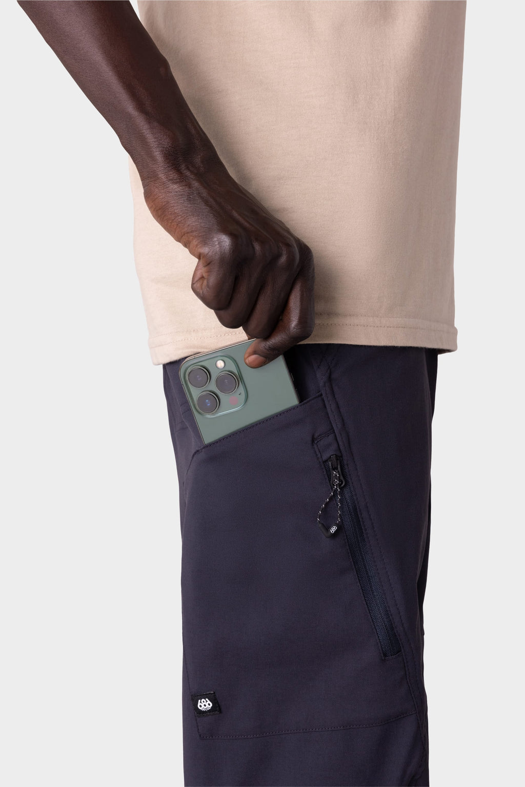 A person in a beige shirt and 686 Men's Anything Cargo Pant - Relaxed Fit by 686 puts a green smartphone with three cameras into the zippered pocket of their 4-way stretch cargo pants.