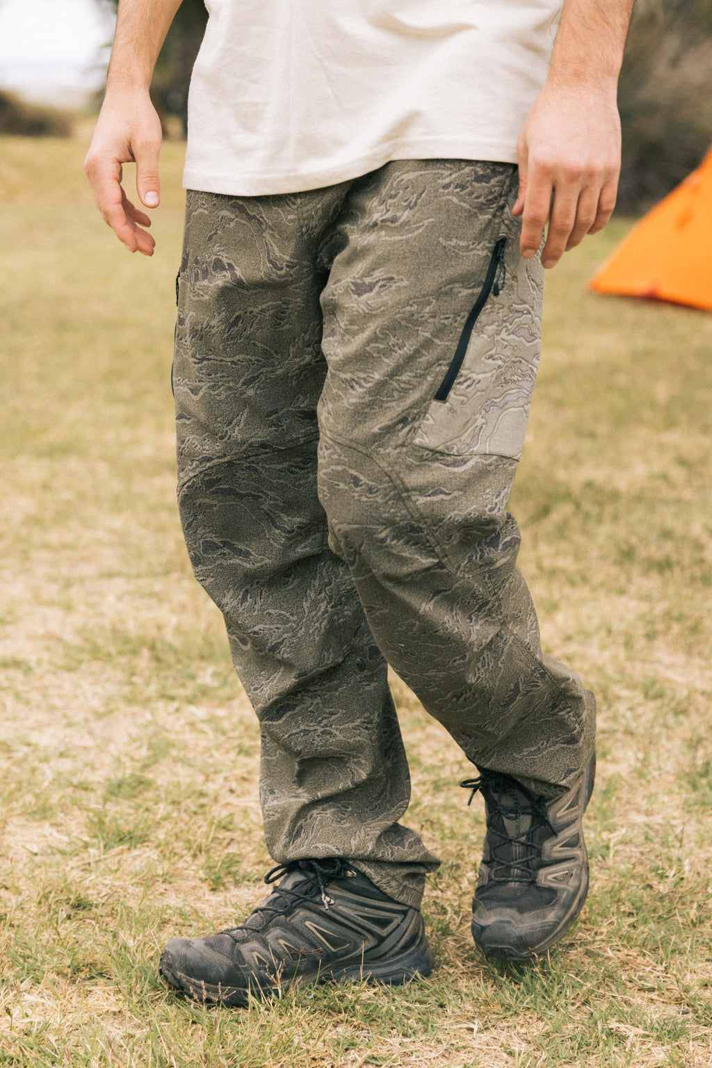 A person stands on grass wearing a white shirt, black hiking shoes, and the 686 Men's Anything Cargo Pant - Relaxed Fit by 686 in olive green. An orange tent appears in the background.