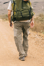 Wearing a large green backpack and 686 Men's Anything Cargo Pant - Relaxed Fit, a person walks along a dirt trail in a dry, natural landscape, seen from behind.